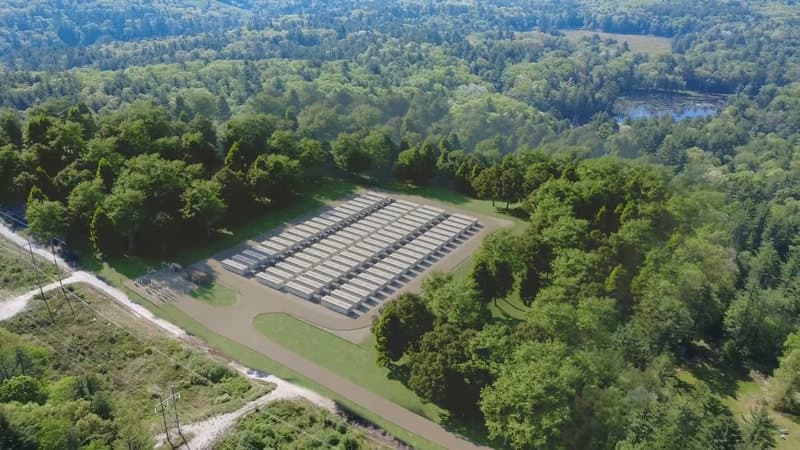 A rendering of the battery storage site as currently designed, seen from above: rows of battery units on a pad in the woods, the project substation at one corner, and the access drive beside the transmission line corridor